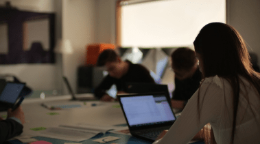 Llun o pobl yn eistedd o gwmpas desg gyda laptops